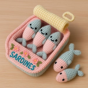 以下が含まれることがあります： ピンク色の手編みのイワシ缶。中には3匹の笑顔の魚と、さらに2匹の魚が入っています。魚はピンクと青で、黒い目があります。缶には青色で「SARDINES」の文字が書かれています。