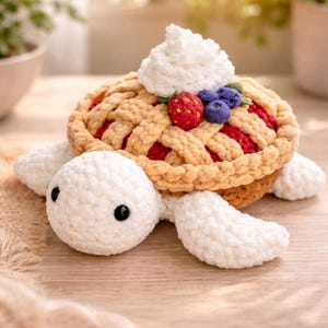 以下が含まれることがあります： パイをテーマにした甲羅を持つ、かぎ針編みのカメの形をしたぬいぐるみ。甲羅は格子模様のタン色で、白いクリーム、赤いイチゴ、青いブルーベリーがトッピングされています。カメは白いヒレと頭を持っています。