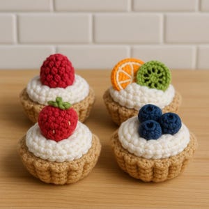 Può includere: Quattro crostate di frutta all'uncinetto. Ogni crostata ha una crosta marrone, un ripieno di crema bianca ed è ricoperta da un frutto diverso: un lampone rosso, una fragola rossa, una fetta di arancia, una fetta di kiwi e tre mirtilli.