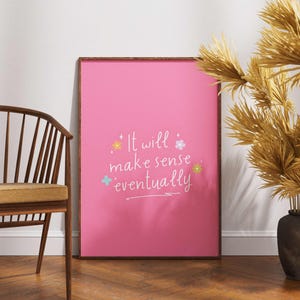 May include: A framed pink poster with the text "It will make sense eventually" in white script, accented with colorful floral and star illustrations. A wooden chair and decorative dried plants in vases are also visible.