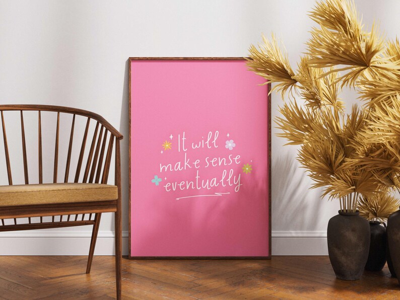 May include: A framed pink poster with the text "It will make sense eventually" in white script, accented with colorful floral and star illustrations. A wooden chair and decorative dried plants in vases are also visible.