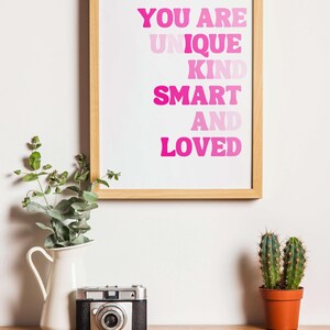 May include: A framed print with the words "YOU ARE UNIQUE KIND SMART AND LOVED" in pink lettering. A vintage camera, a white pitcher with greenery, and a small potted cactus sit on a wooden surface.