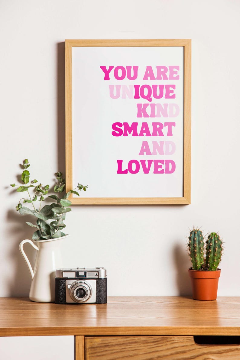 May include: A framed print with the words "YOU ARE UNIQUE KIND SMART AND LOVED" in pink lettering. A vintage camera, a white pitcher with greenery, and a small potted cactus sit on a wooden surface.
