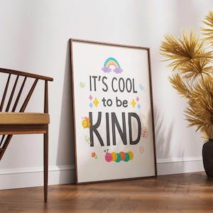 May include: A framed poster with the words "It's cool to be KIND" in a colorful design. The poster features a rainbow, stars, a bee, and a caterpillar. The frame is brown, and the poster is leaning against a white wall.