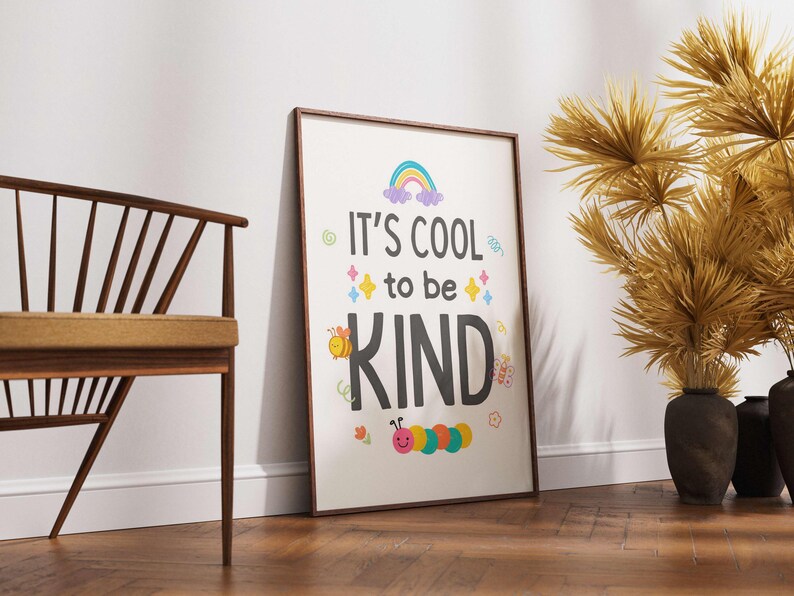 May include: A framed poster with the words "It's cool to be KIND" in a colorful design. The poster features a rainbow, stars, a bee, and a caterpillar. The frame is brown, and the poster is leaning against a white wall.