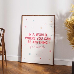 May include: A framed print with the text "IN A WORLD WHERE YOU CAN BE ANYTHING be kind" in pink and light pink. The print has colorful star accents. The frame is brown, and the print is leaning against a white wall.