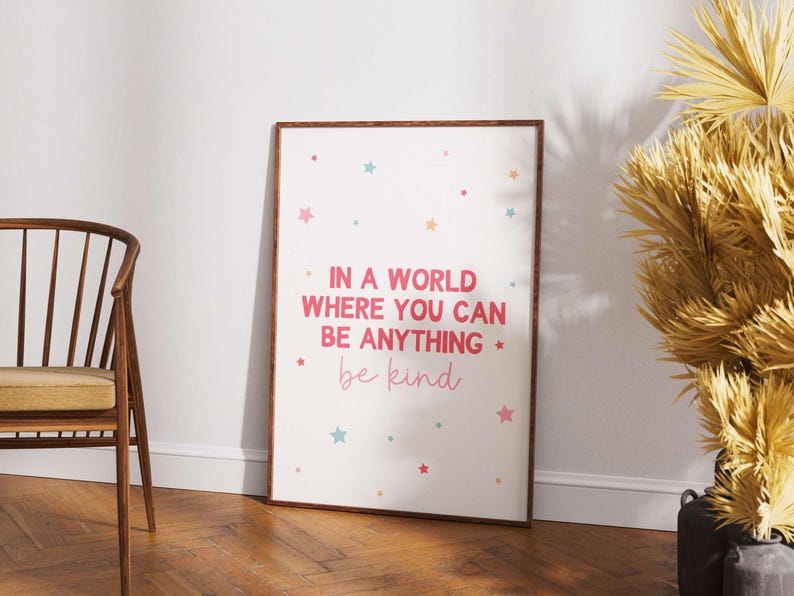 May include: A framed print with the text "IN A WORLD WHERE YOU CAN BE ANYTHING be kind" in pink and light pink. The print has colorful star accents. The frame is brown, and the print is leaning against a white wall.