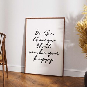 May include: A framed print with the text "Do the things that make you happy" in black script. The frame is brown, and the print is leaning against a white wall. A wooden chair and decorative dried plants are also in the image.