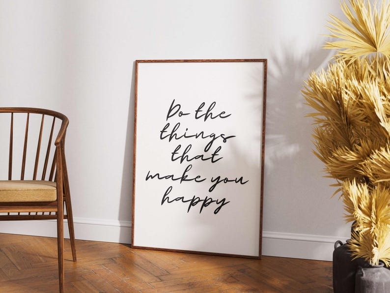 May include: A framed print with the text "Do the things that make you happy" in black script. The frame is brown, and the print is leaning against a white wall. A wooden chair and decorative dried plants are also in the image.