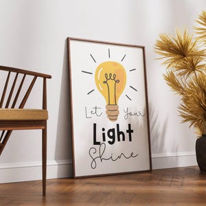 May include: A framed print with a yellow lightbulb illustration and the words "Let Your Light Shine" in black and script font. The artwork is leaning against a white wall on a wooden floor. A wooden chair and decorative plants are also in the image.
