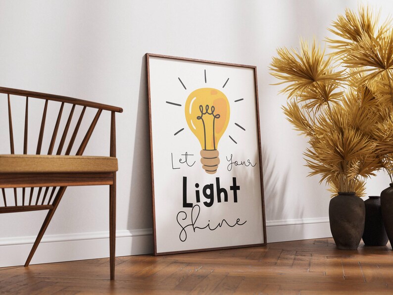 May include: A framed print with a yellow lightbulb illustration and the words "Let Your Light Shine" in black and script font. The artwork is leaning against a white wall on a wooden floor. A wooden chair and decorative plants are also in the image.
