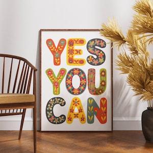 May include: A framed art print with the words "YES YOU CAN" in colorful, floral-patterned letters. The letters are in shades of red, yellow, green, and blue. The print is displayed against a white wall, with a wooden chair and decorative vase in the foreground.
