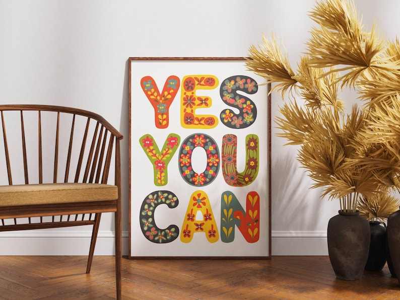 May include: A framed art print with the words "YES YOU CAN" in colorful, floral-patterned letters. The letters are in shades of red, yellow, green, and blue. The print is displayed against a white wall, with a wooden chair and decorative vase in the foreground.
