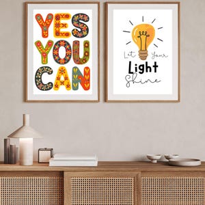 May include: Two framed art prints. One print says "YES YOU CAN" in colorful, floral-patterned letters. The other print features a yellow lightbulb with the words "Let Your Light Shine". Both prints are in wooden frames.