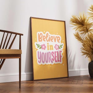 May include: A framed poster with the words "Believe in Yourself" in a colorful, playful font. The poster is set against a yellow background and decorated with floral accents. The frame is brown, and the poster is leaning against a white wall.