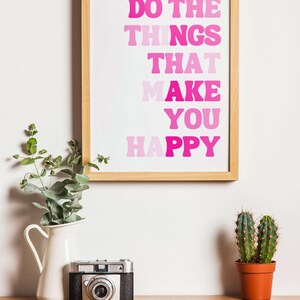 May include: A framed print with the text "DO THE THINGS THAT MAKE YOU HAPPY" in shades of pink. A vintage camera, a white pitcher with greenery, and a small cactus in a terracotta pot sit on a wooden surface.