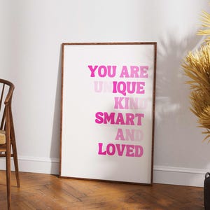 May include: A framed art print with the words "YOU ARE UNIQUE KIND SMART AND LOVED" in shades of pink. The print is displayed on a white background, with a wooden frame. A wooden chair and decorative plant are in the background.