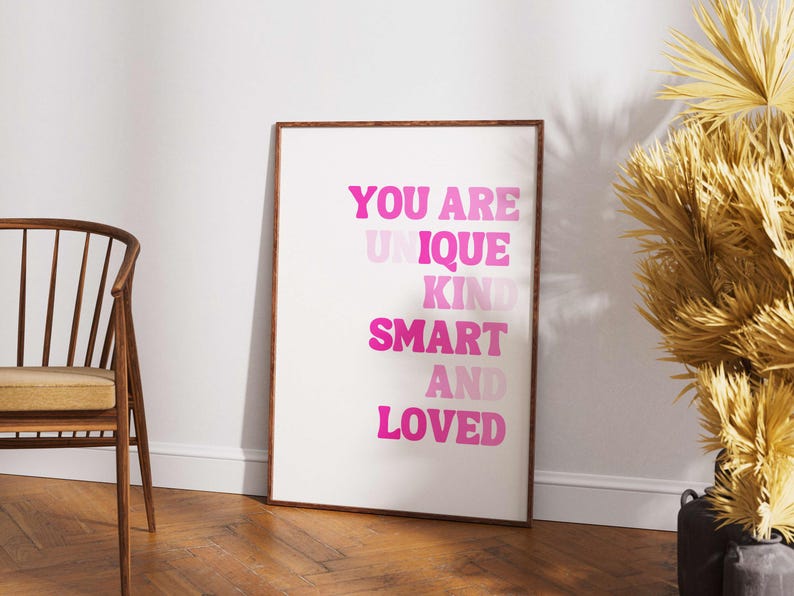 May include: A framed art print with the words "YOU ARE UNIQUE KIND SMART AND LOVED" in shades of pink. The print is displayed on a white background, with a wooden frame. A wooden chair and decorative plant are in the background.