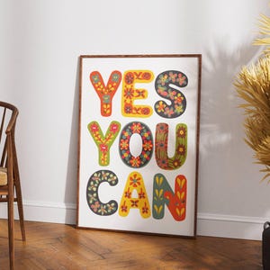 May include: A framed art print with the words "YES YOU CAN" in large, colorful, floral-patterned letters. The artwork is displayed on a white wall, with a wooden chair and a decorative plant in the background.
