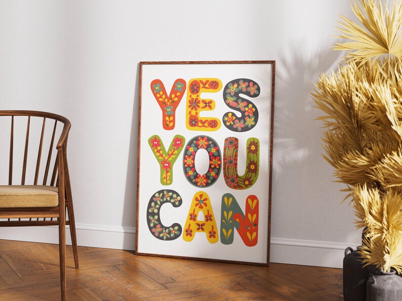 May include: A framed art print with the words "YES YOU CAN" in large, colorful, floral-patterned letters. The artwork is displayed on a white wall, with a wooden chair and a decorative plant in the background.