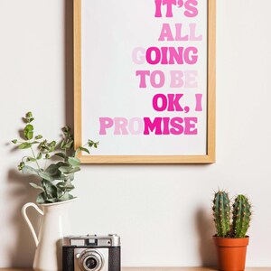 May include: A framed print with the text "IT'S ALL GOING TO BE OK, I PROMISE" in shades of pink. A white pitcher with greenery, a vintage camera, and a potted cactus sit on a wooden surface.