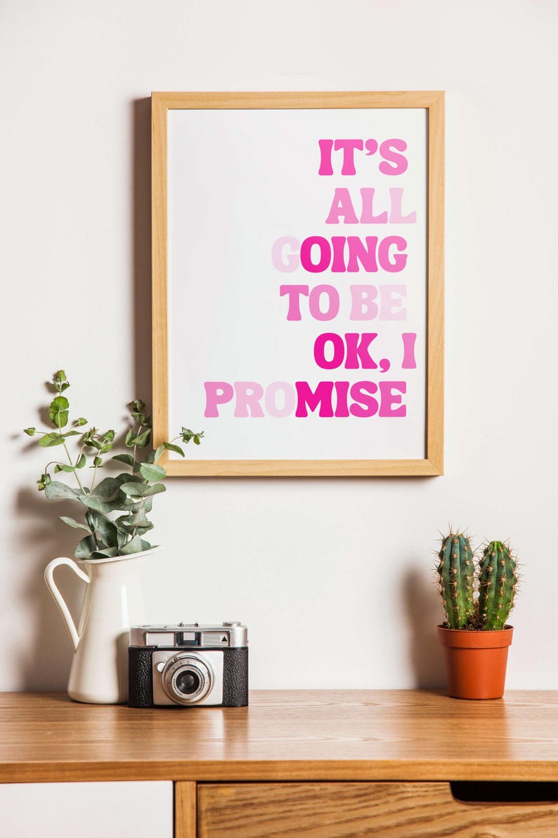 May include: A framed print with the text "IT'S ALL GOING TO BE OK, I PROMISE" in shades of pink. A white pitcher with greenery, a vintage camera, and a potted cactus sit on a wooden surface.