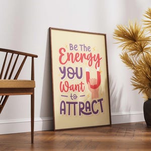 May include: Framed poster with the text "Be The Energy You Want to ATTRACT" in red, purple, and white. The poster is leaning against a white wall on a wooden floor. A wooden chair and decorative dried plants are also in the image.