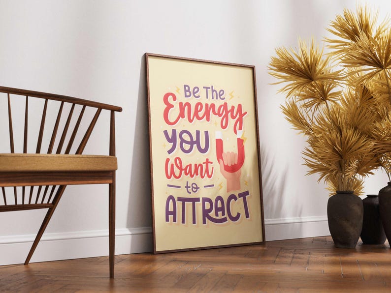 May include: Framed poster with the text "Be The Energy You Want to ATTRACT" in red, purple, and white. The poster is leaning against a white wall on a wooden floor. A wooden chair and decorative dried plants are also in the image.
