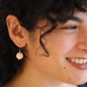 Peut inclure: Gros plan sur une oreille portant une boucle d'oreille dorée. La boucle d'oreille est ornée d'une perle ronde blanc cassé suspendue à un crochet doré. La personne sourit, avec des cheveux bouclés foncés encadrant son visage.