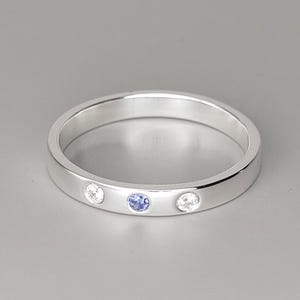 Può includere: Un anello d'argento con tre pietre preziose. L'anello presenta una gemma blu centrale affiancata da due gemme trasparenti. L'anello è posto su uno sfondo grigio.