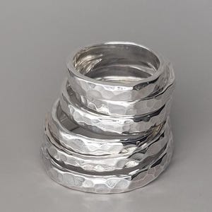 May include: A stack of silver rings with a hammered texture. The rings are round and have a slightly irregular surface, reflecting light. The rings are stacked on top of each other, creating a layered effect. The background is a solid grey.