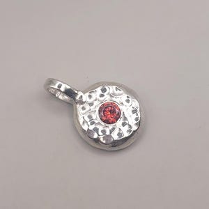 Puede incluir: Un colgante circular de plata con una superficie texturizada y martillada, que presenta una pequeña piedra preciosa roja, redonda y facetada, engastada en el centro. El colgante incluye una anilla de plata en la parte superior para una cadena.
