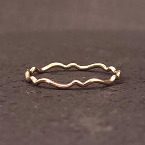 May include: A thin, delicate ring crafted from a polished rose gold-toned metal. The band features a continuous, gentle wavy or undulating design that creates a soft, organic silhouette. The surface of the metal is smooth and reflective.