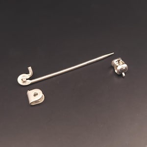 Op de afbeelding: Een zilverkleurige hoedenspeld met een lange, rechte schacht en een puntig uiteinde. De speld heeft een decoratieve kop en een aparte sluiting. De onderdelen worden tegen een donkere achtergrond getoond, wat het ontwerp en de details van de speld benadrukt.