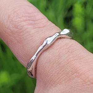 Op de afbeelding: Een zilveren ring met een organisch, getextureerd ontwerp. De ring wordt aan een vinger gedragen, met een vage groene achtergrond. Het oppervlak van de ring reflecteert licht, wat de onregelmatige vorm benadrukt. Het sieraad lijkt handgemaakt.
