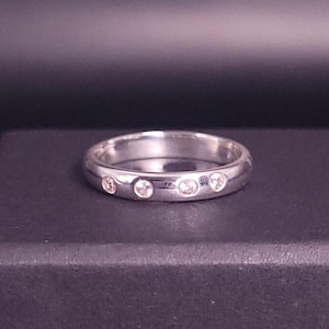 Può includere: Un anello d'argento con tre piccole pietre preziose trasparenti. L'anello è appoggiato su una scatola nera, con uno sfondo sfumato scuro. L'anello sembra essere in metallo lucido.
