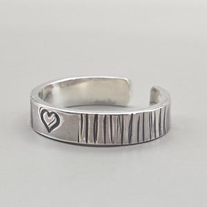 Op de afbeelding: Een zilverkleurige ring met een hartvormig ontwerp. De ring heeft een getextureerde band met verticale lijnen en een open ontwerp. Het hart is in het metaal geëtst. De ring wordt getoond op een grijze achtergrond.