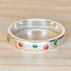 Può includere: Un anello d'argento con una serie di pietre preziose arancioni e verdi alternate. L'anello è incastonato su uno sfondo chiaro di venature del legno, evidenziando il metallo lucido e le pietre colorate. L'anello è uno stile a fascia.