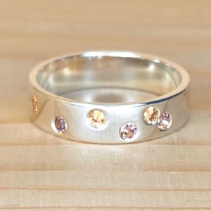 May include: A silver ring with a flat band, adorned with several small, round gemstones in shades of peach and purple. The ring is set against a light wooden surface, highlighting its polished metal and colorful accents.
