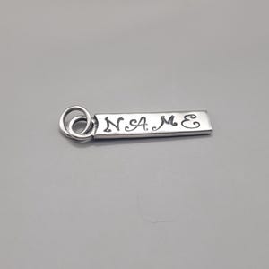May include: A silver-colored rectangular keychain tag with the word "NAME" engraved in a cursive font. The tag has a small loop at one end for attaching to a keyring. The background is a solid gray.