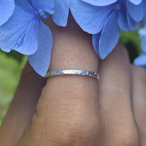 Puede incluir: Un anillo de plata con textura martillada se muestra en un dedo, sobre un fondo de vibrantes flores de hortensia azul. La superficie del anillo refleja la luz, mostrando su diseño único. El anillo es una delicada pieza de joyería.