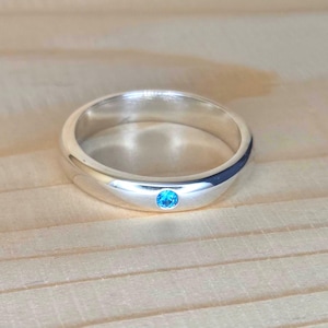 Könnte beinhalten: Ein silberner Ring mit einem einzelnen, runden, hellblauen Edelstein in der Mitte. Der Ring hat eine glatte, polierte Oberfläche und liegt auf einer hellen Holzoberfläche.