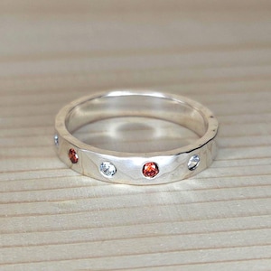 May include: Silver ring with a hammered texture, featuring alternating clear and red gemstones. The ring is set against a light wood grain background, showcasing the jewelry's design and craftsmanship.