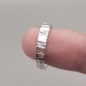 May include: A silver ring with a textured, rectangular design. The ring is held by a finger, showcasing its polished surface and geometric pattern. The ring's band is approximately 0.25 inches wide.