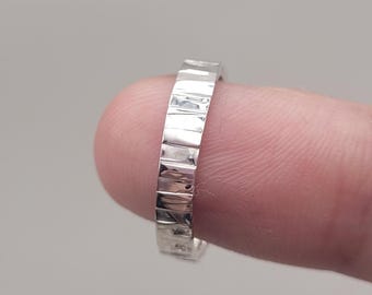 May include: A silver ring with a textured, rectangular design. The ring is held by a finger, showcasing its polished surface and geometric pattern. The ring's band is approximately 0.25 inches wide.