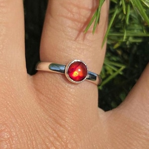 Op de afbeelding: Een zilveren ring met een ronde, rode edelsteen. De edelsteen heeft een vurige, meerkleurige uitstraling. De ring zit om een vinger, met groen gebladerte op de achtergrond.