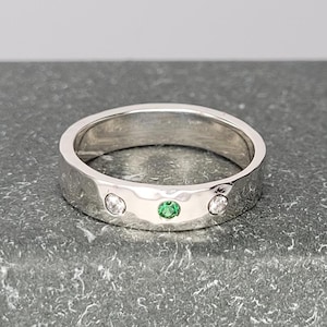 May include: A silver ring with three gemstones. The center stone is green, flanked by two clear stones. The ring is set on a gray surface.