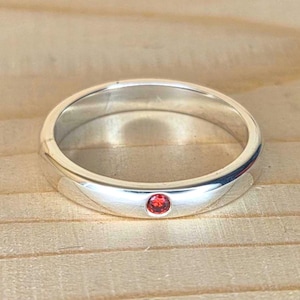 Puede incluir: Un anillo de plata con una pequeña gema roja redonda engastada en el centro. El anillo está sobre una superficie de madera, y la banda tiene un acabado liso y pulido. La gema es de color rojo vibrante.