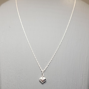 Op de afbeelding: Een zilverkleurige ketting met een hartvormige hanger. De hanger is klein en afgerond, met een glad oppervlak. De ketting is een delicate, gedraaide touwstijl. De achtergrond is effen, gedempt grijs.
