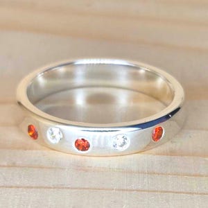 Può includere: Un anello d'argento con finitura lucida, caratterizzato da una serie di pietre preziose rosse e chiare alternate. L'anello è posto su uno sfondo chiaro con venature del legno, che mette in risalto il design e la maestria del gioiello.